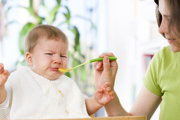 Crying baby boy refusing to eat food from spoon with hands dirty